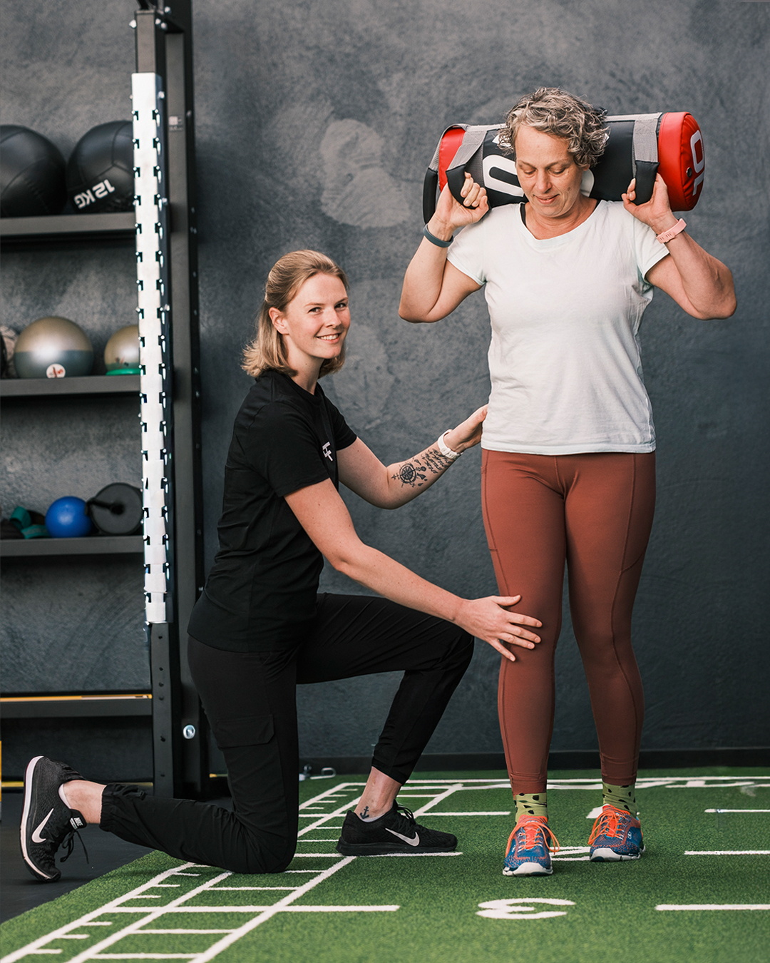 Eine Trainerin unterstützt eine Frau beim Training mit einem Gewichtssack auf den Schultern