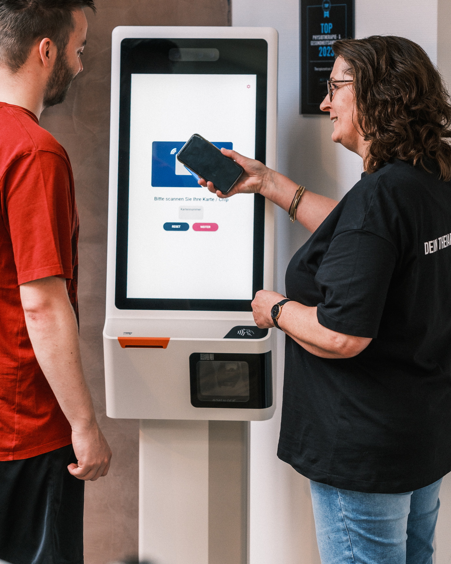 Eine Mitarbeiterin erklärt einem Patienten die Nutzung eines digitalen Terminals zur Anmeldung